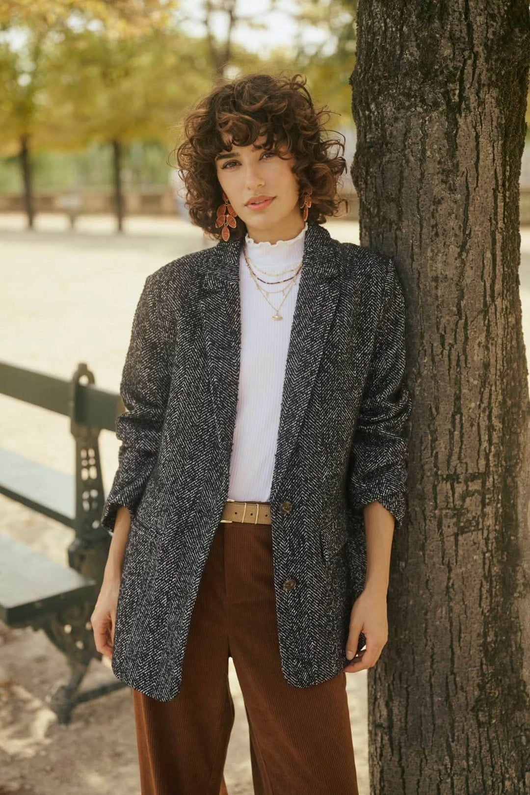Person wearing a tweed blazer with a white t-shirt and brown trousers standing by a tree in a park setting.