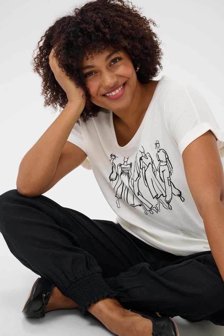 Femme souriante portant un T-shirt Rigga en coton avec imprimé graphique, assise en pantalon noir.