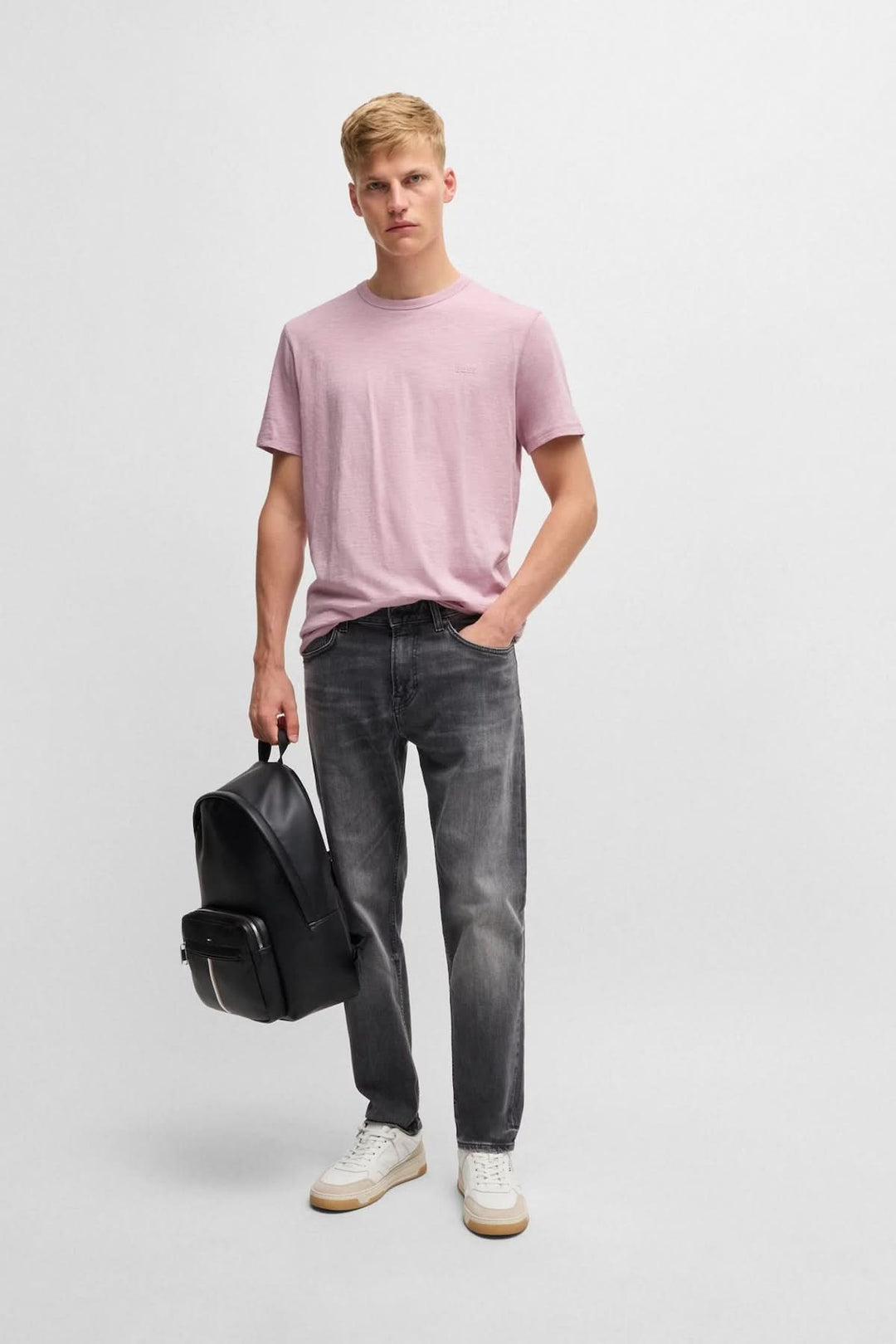 Homme portant un t-shirt rose avec logo, jeans gris et sac à dos noir, style décontracté.