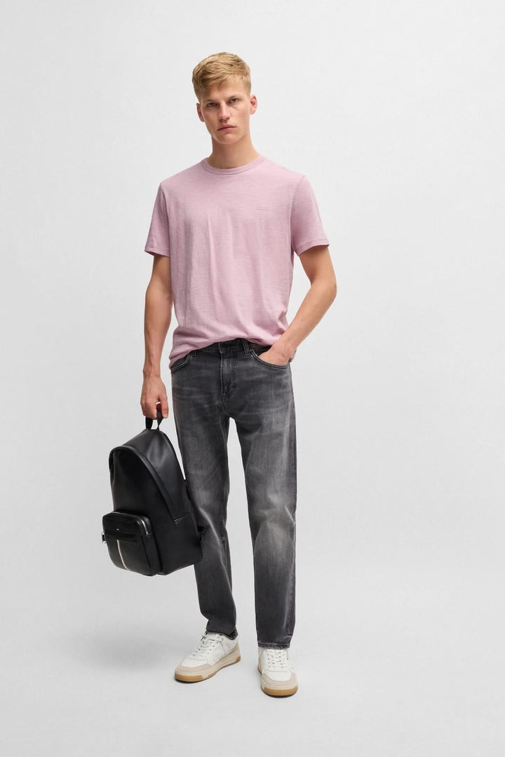 Homme portant un t-shirt rose avec logo, jeans gris et sac à dos noir, style décontracté.