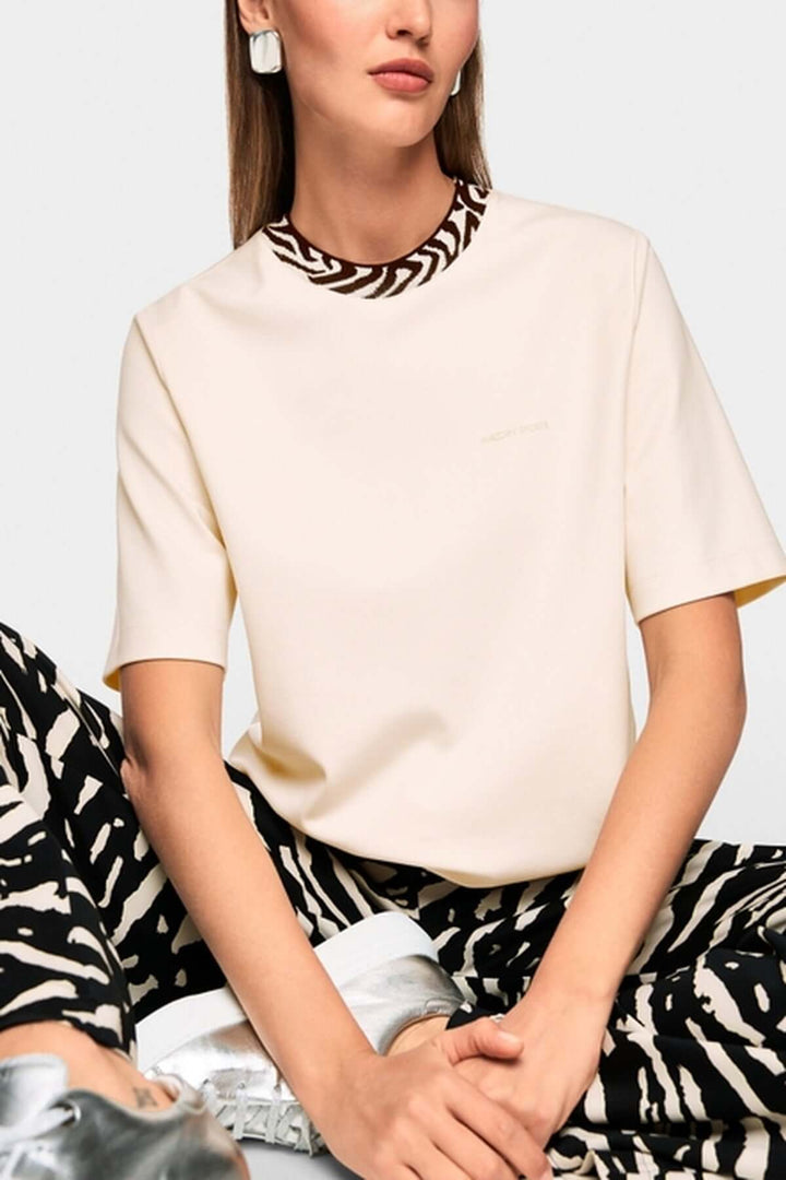 T-shirt classique avec col contrasté et logo Marc Cain Sport, coupe droite et manches courtes, porté par un mannequin assis.