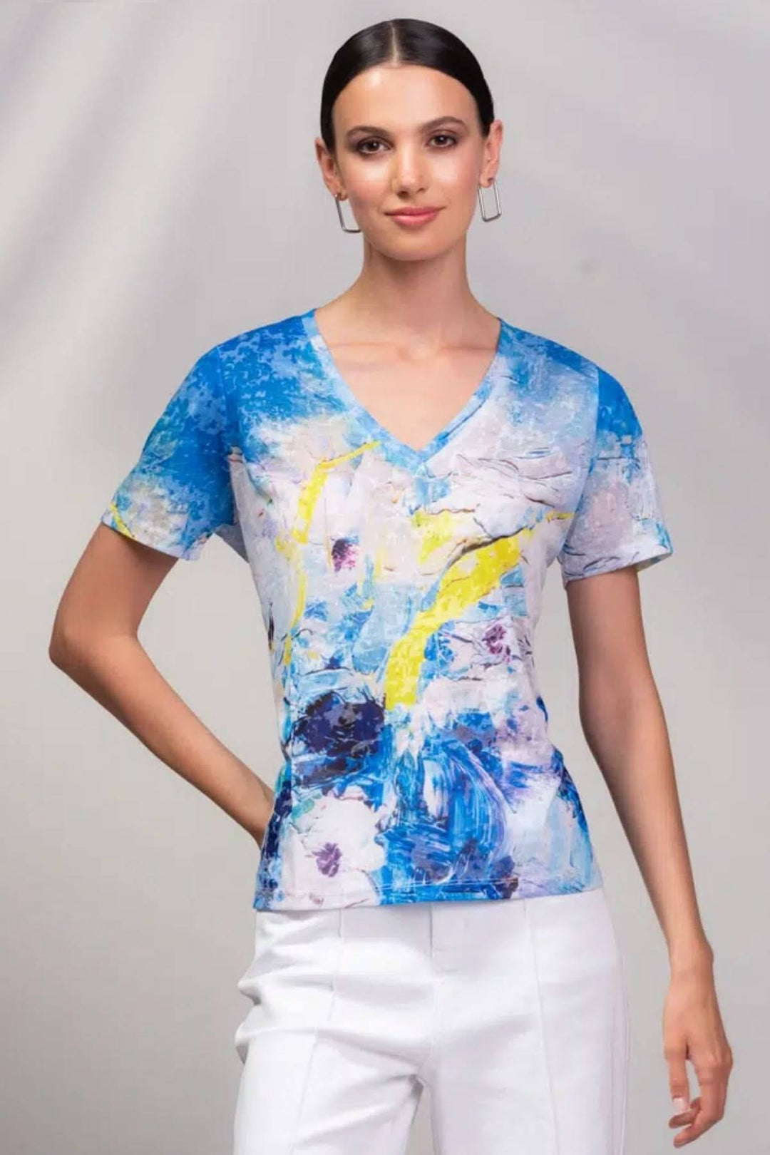 T-shirt à col en V, imprimé artistique façon aquarelle en bleu et jaune, porté avec un pantalon blanc.