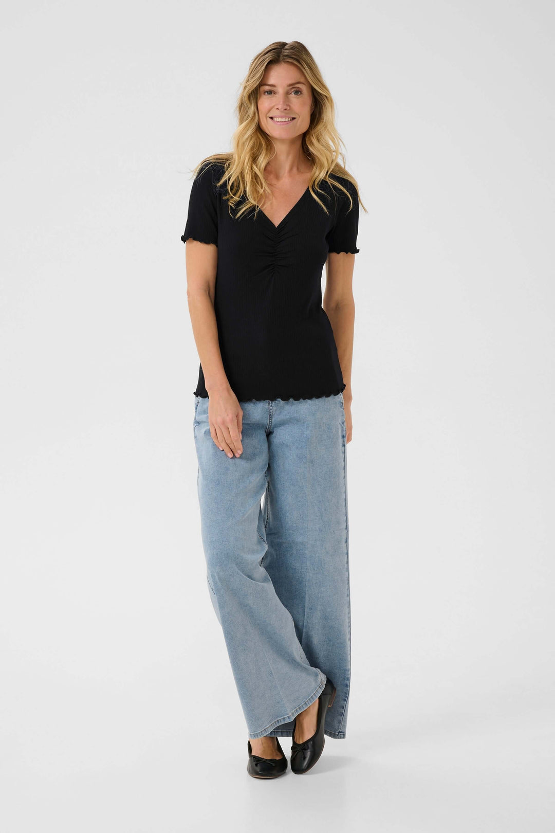 Femme portant un t-shirt noir à col froncé et un pantalon large en jean, souriante sur fond blanc.