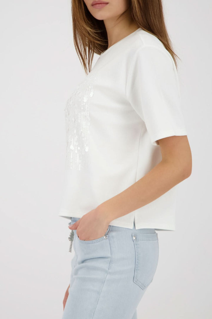 Femme portant un T-shirt à empiècement pailleté, coupe droite, encolure ronde, avec un jean clair, style moderne et féminin.
