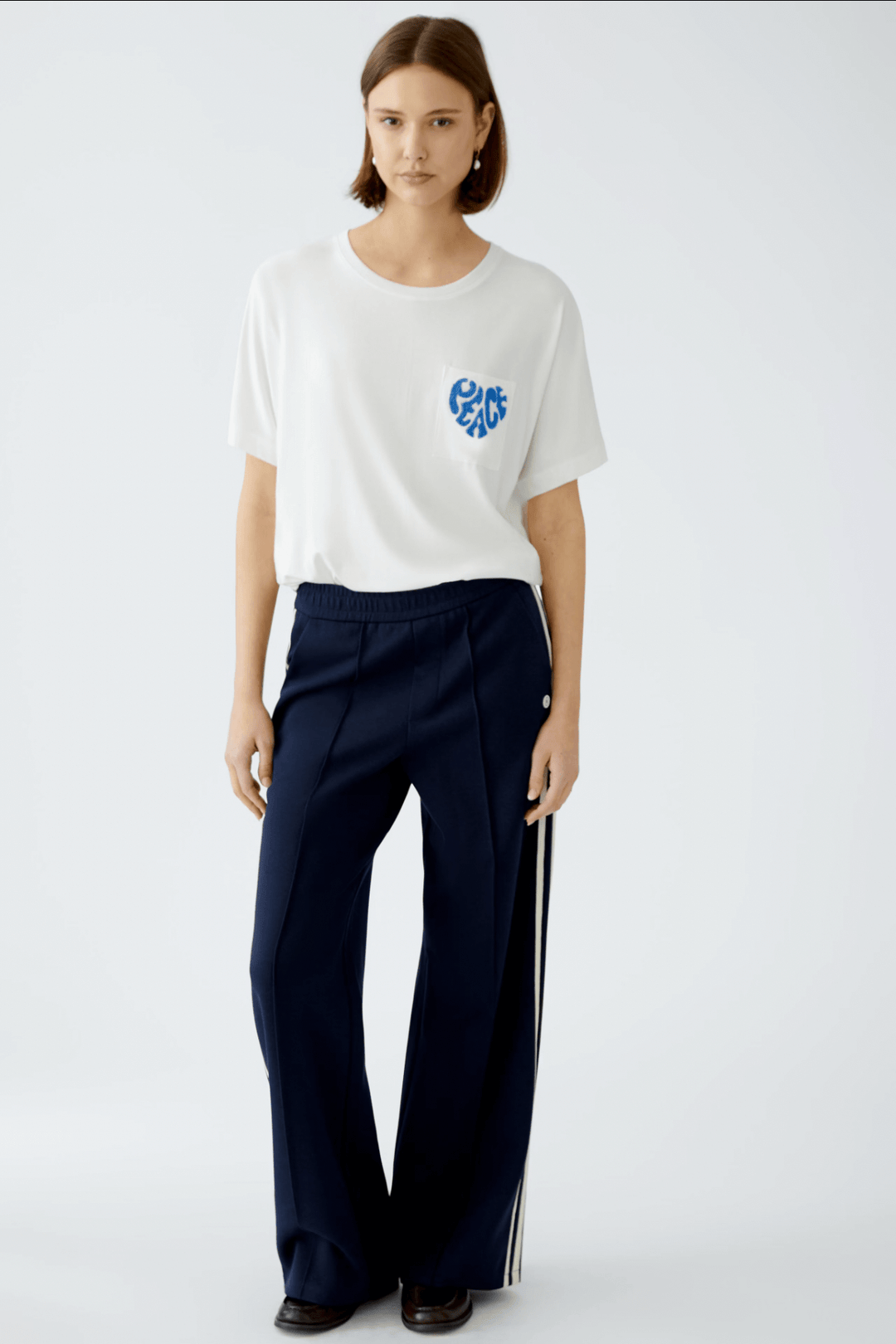 Femme portant un t-shirt blanc avec broderie "Peace" sur la poche, associé à un pantalon bleu.