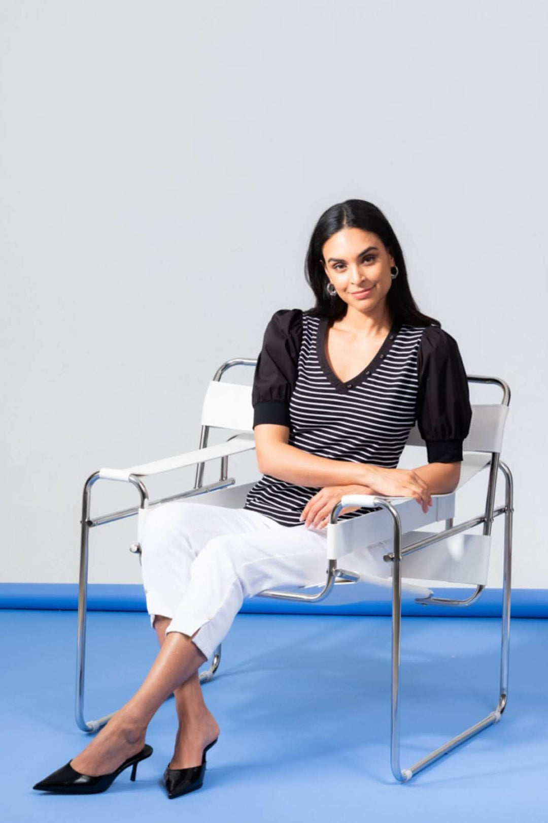 Femme portant un t-shirt à rayures noires et blanches, assise sur une chaise moderne avec un look chic.