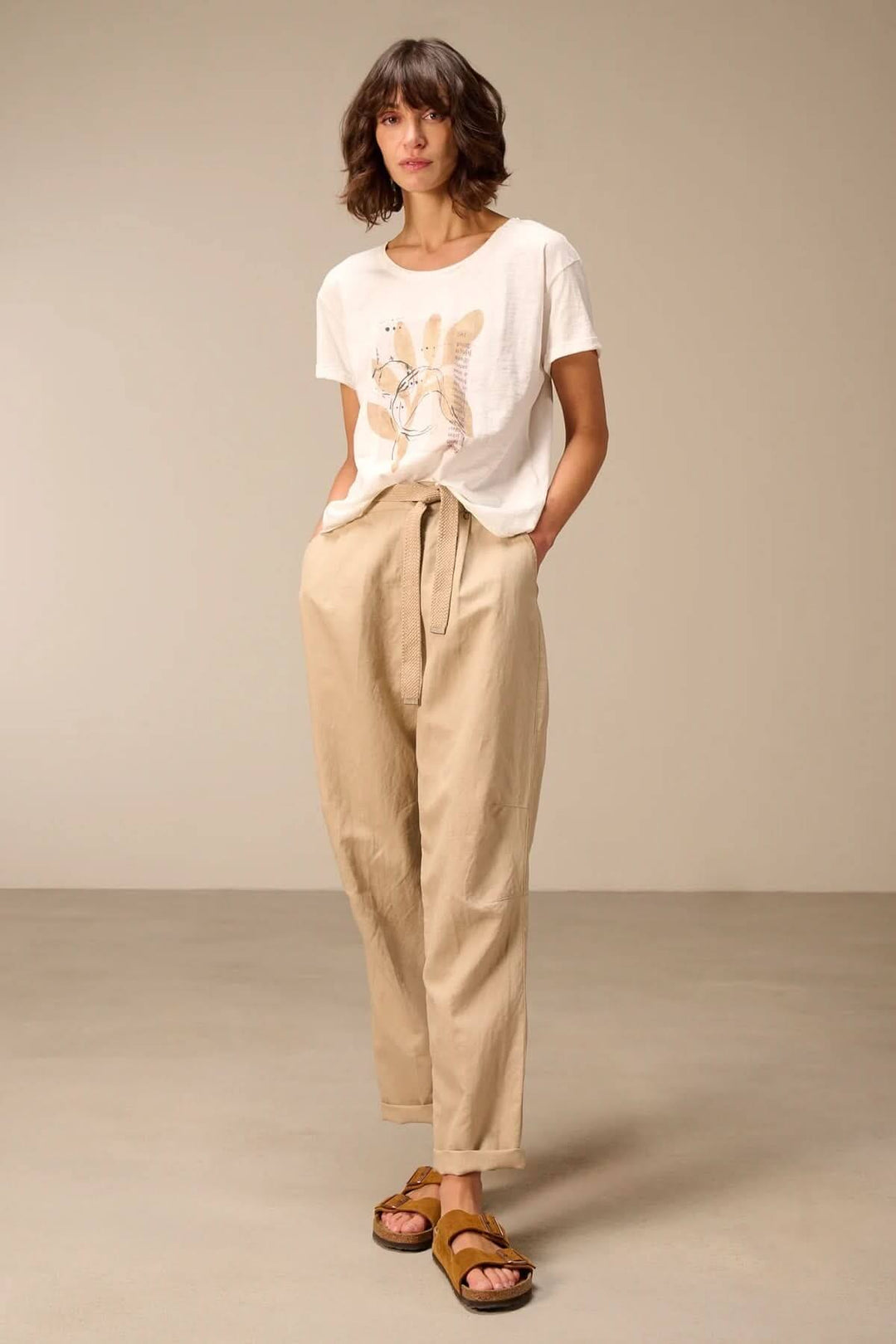 Femme portant un T-shirt à manches courtes coupe décontractée blanc avec un imprimé, assorti à un pantalon beige et des sandales.