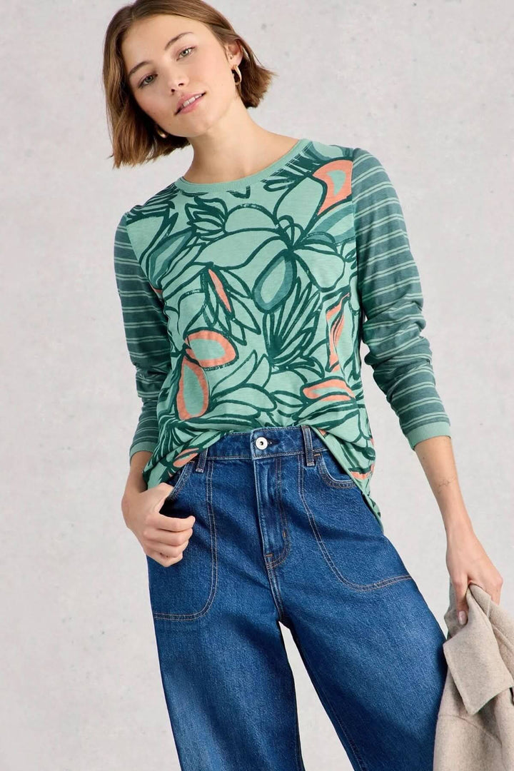 Femme portant le T-shirt à manches longues Clara, avec un motif floral et rayé vert, assorti à un jean. Simple et confortable, idéal au quotidien.