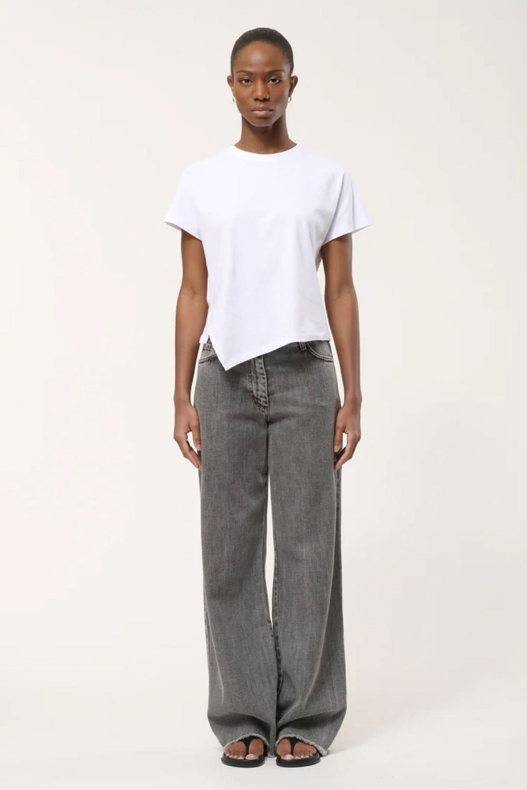 Femme portant un t-shirt à ourlet asymétrique blanc, coupe droite et fluide, avec un pantalon large gris pour un style moderne.
