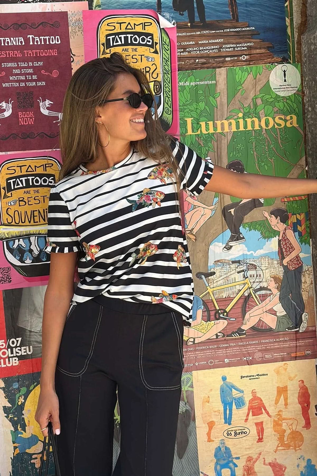 Femme portant un T-shirt rayé avec poissons volants devant un mur coloré de posters vintage.