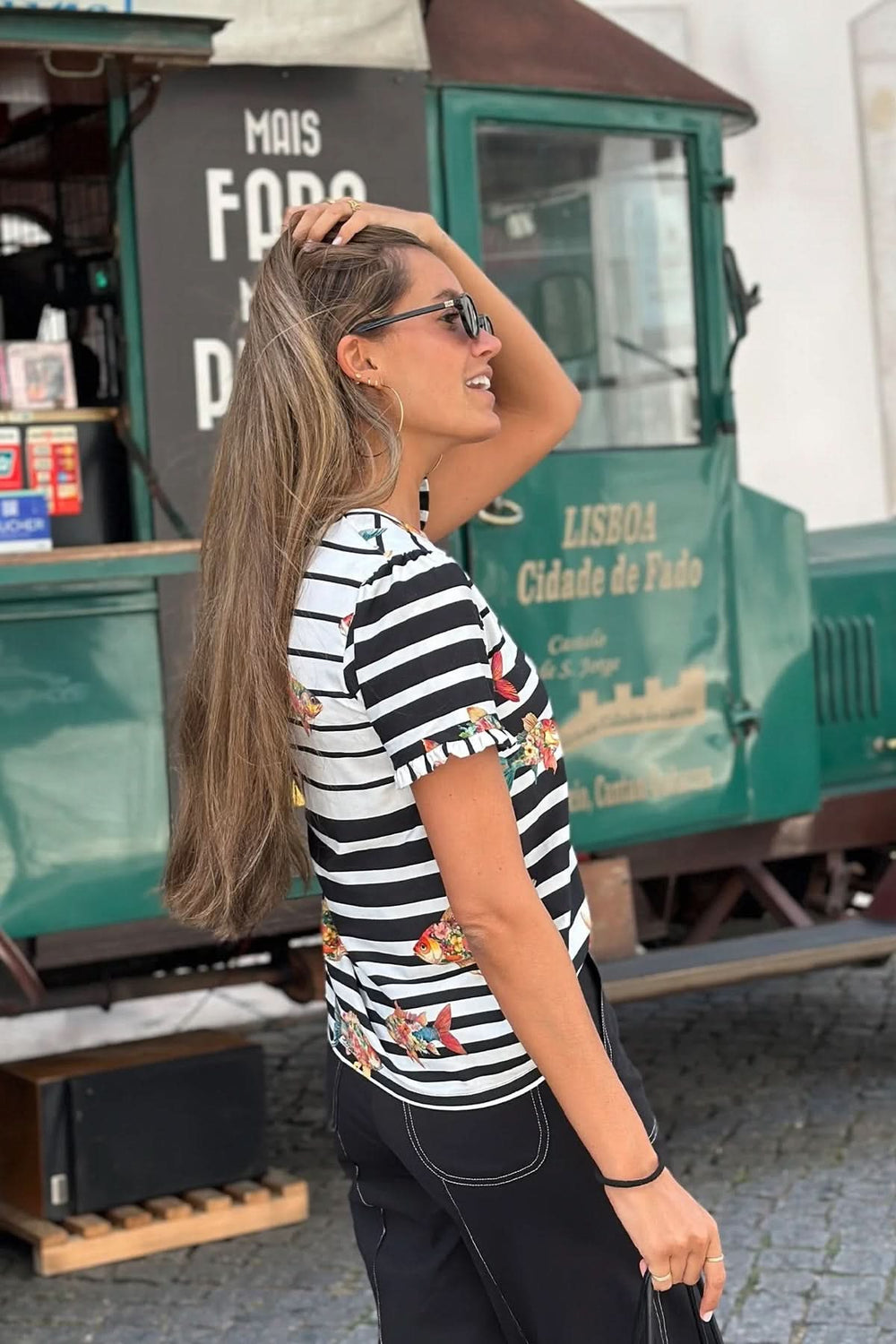Femme portant un t-shirt rayé avec poissons volants, debout devant un camion de nourriture vert à Lisbonne.