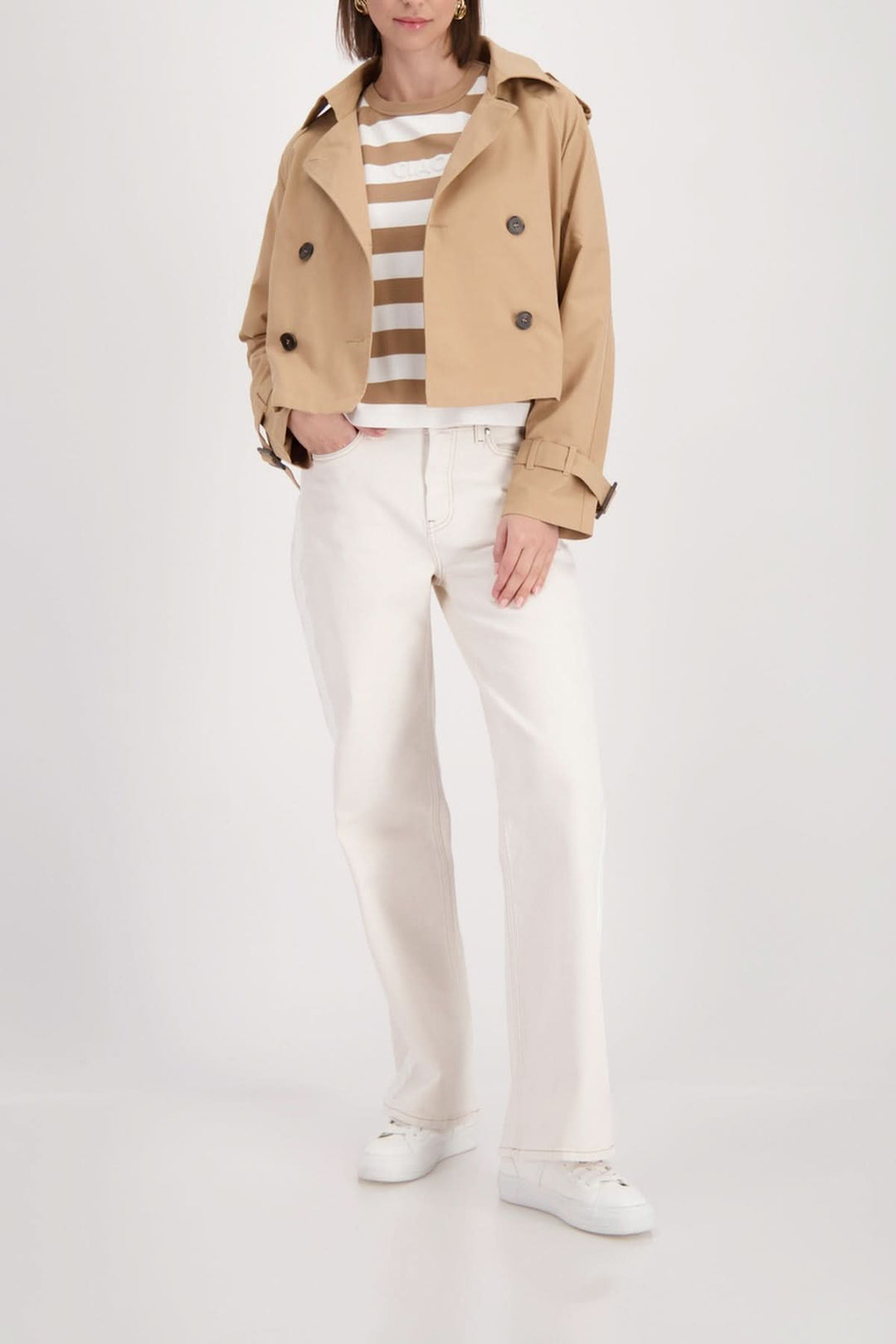 Femme portant un T-shirt rayé à logo embossé sous une veste beige, pantalons blancs et chaussures blanches, style casual chic.