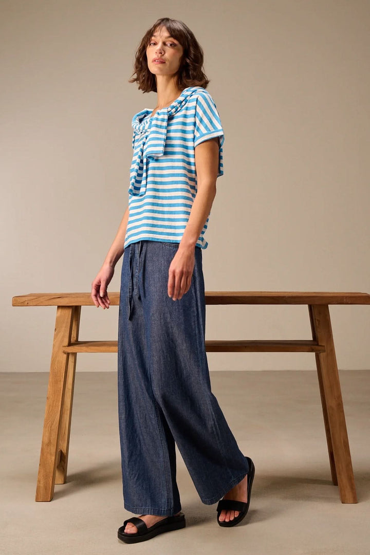 Femme portant un T-shirt rayé à manches courtes coupe décontractée avec un pantalon ample, devant une table en bois.