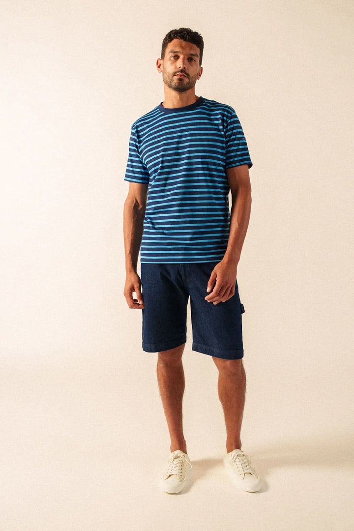 Homme portant un T-shirt à rayures classique bleu et noir avec short, expression décontractée, sur fond beige.