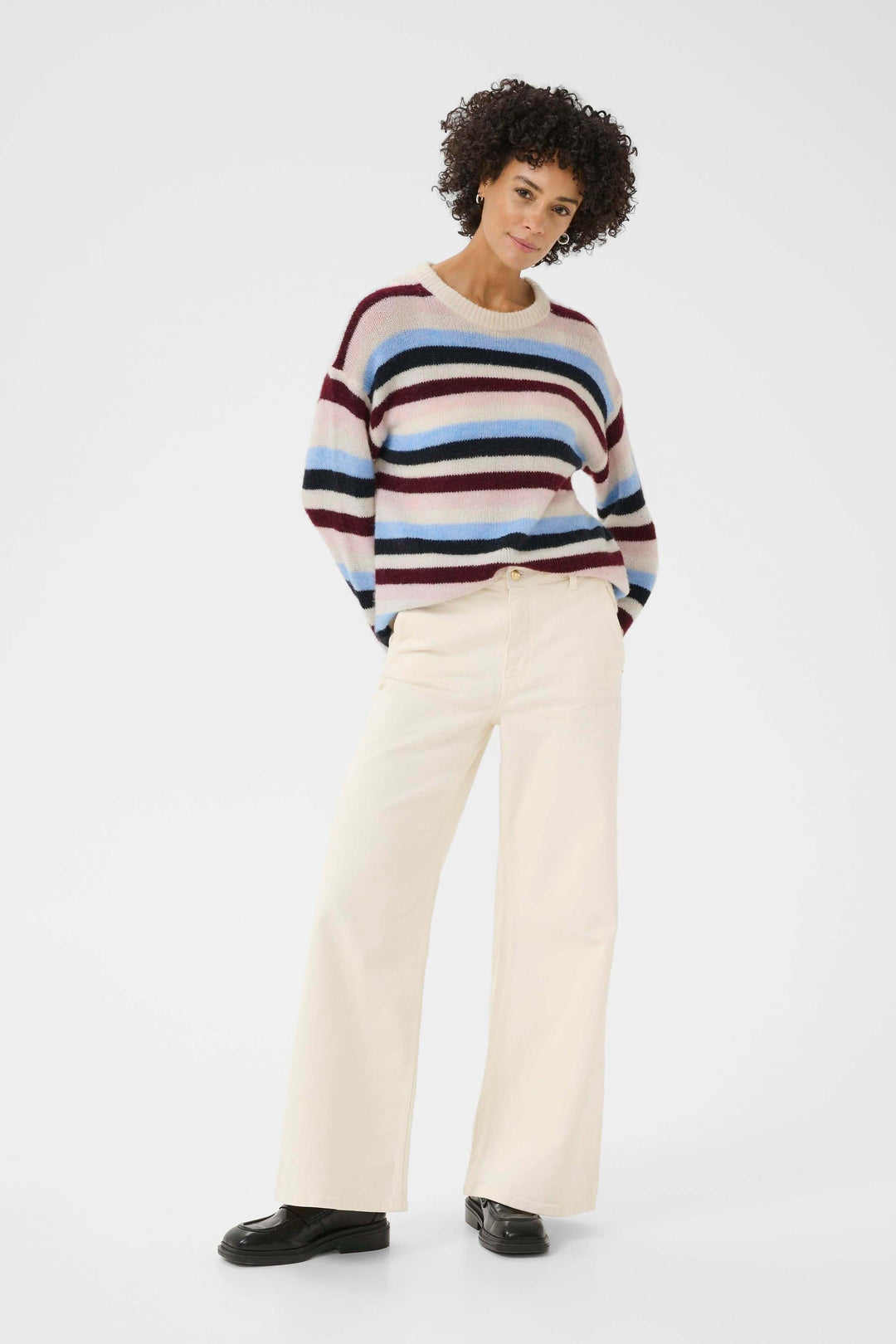 Femme portant un pull rayé coloré avec un pantalon blanc, posant debout. Tricot Spence décontracté et confortable pour un look chic.