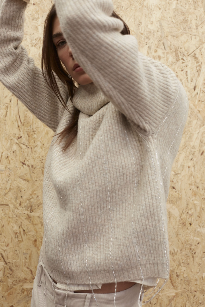 Tricot beige à col roulé avec fils pailletés et broderies scintillantes sur l'ourlet.