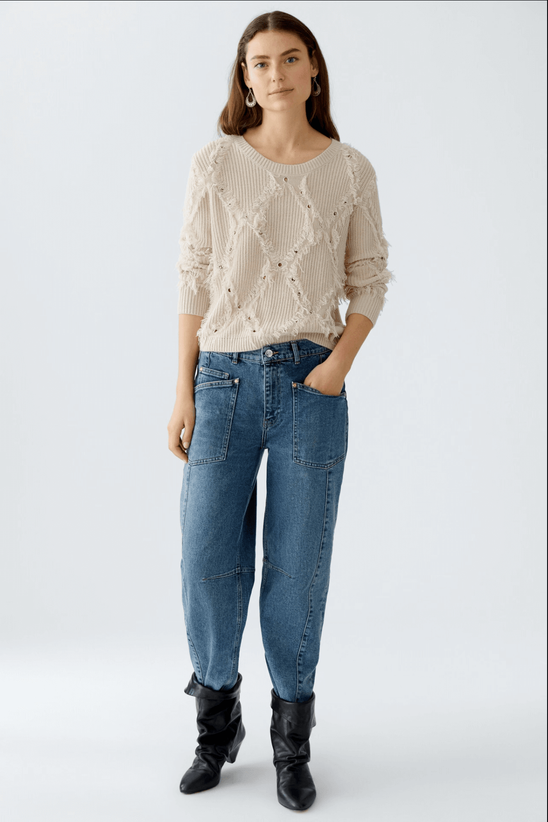 Pull en tricot motif losange avec franges, associé à un pantalon en jean décontracté et bottes noires.