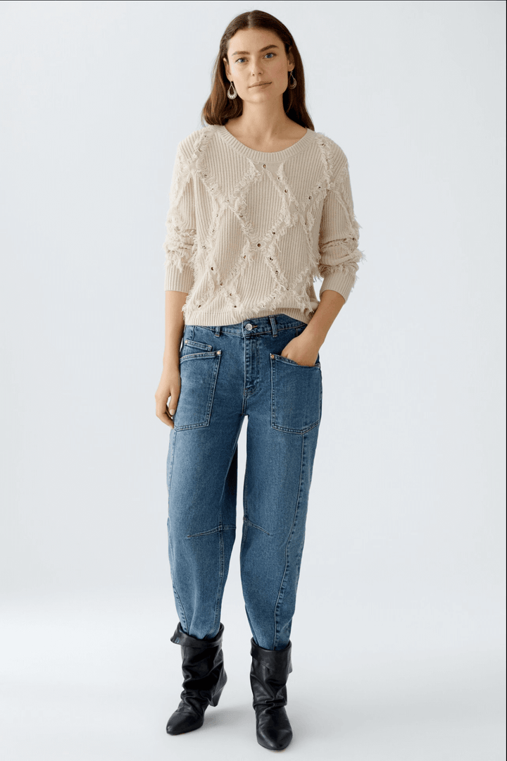 Pull en tricot motif losange avec franges, associé à un pantalon en jean décontracté et bottes noires.