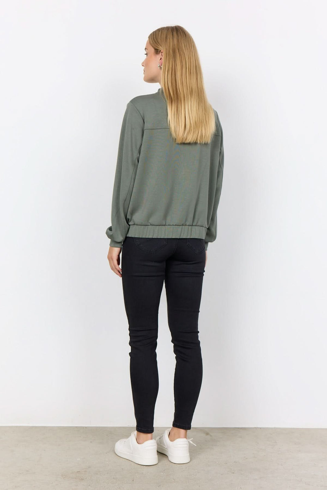 Femme portant une veste à bouton-pression verte, vue arrière, avec des pantalons noirs et des baskets blanches.