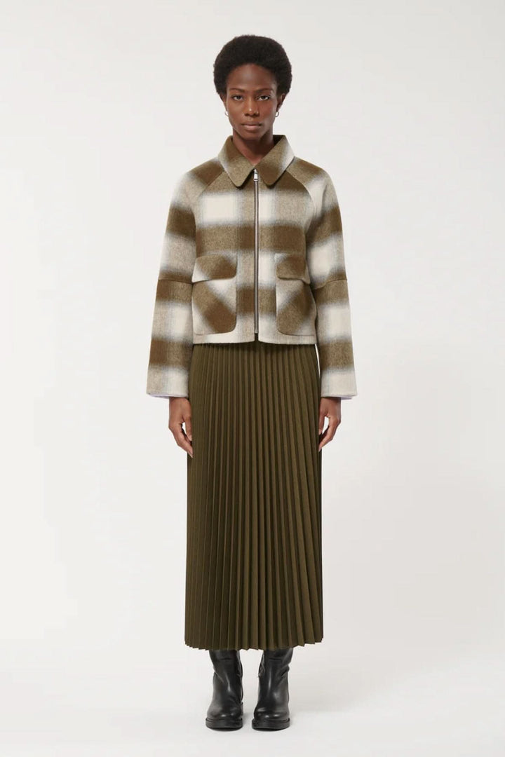 Femme portant une veste courte à carreaux avec poches à rabat et fermeture éclair, associée à une jupe plissée et des bottes noires.
