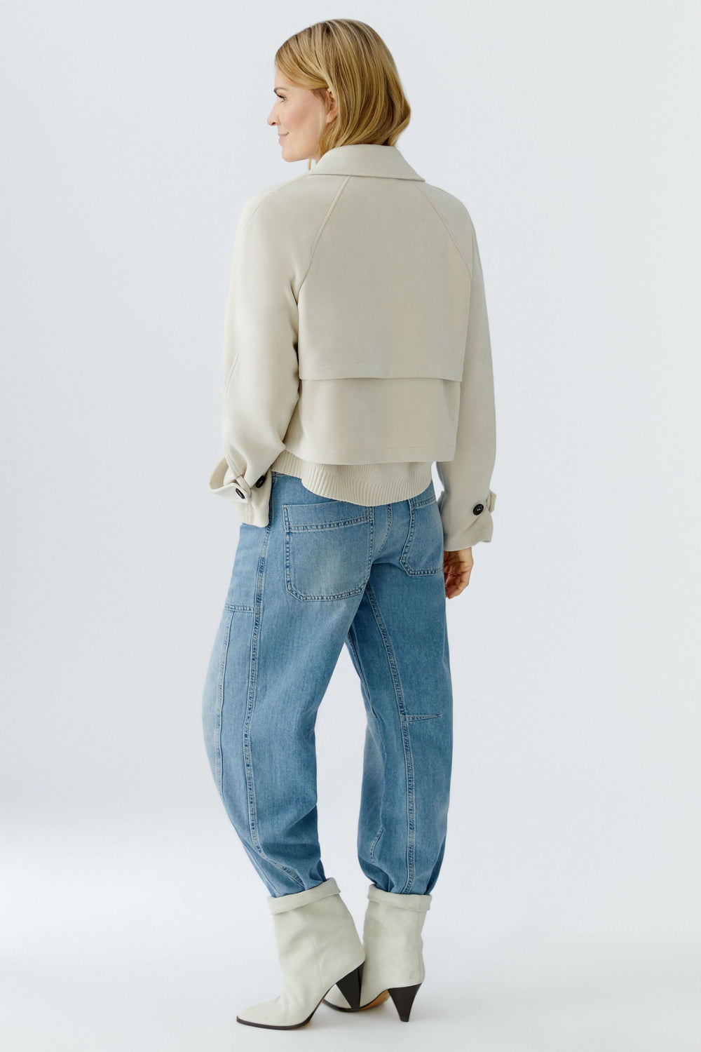 Femme portant une veste courte croisée à col tailleur, vue de dos, stylisée avec un jean et des bottes blanches pour un look moderne.