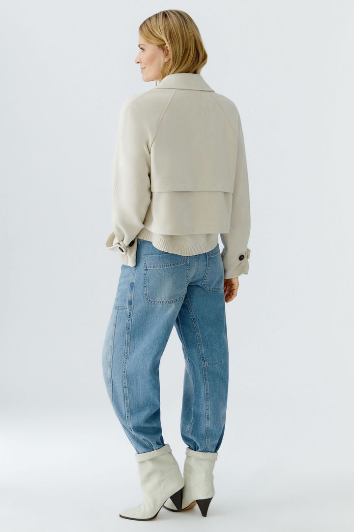 Femme portant une veste courte croisée à col tailleur, vue de dos, stylisée avec un jean et des bottes blanches pour un look moderne.