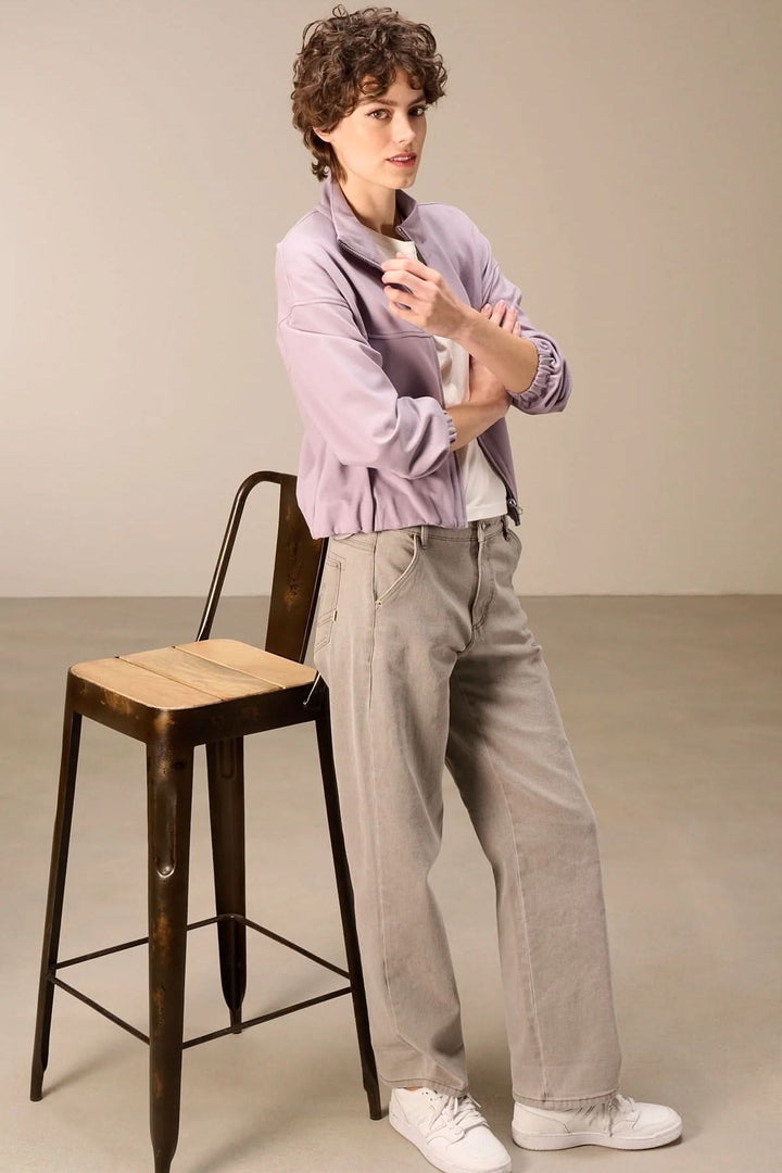 Femme portant une veste en sweat avec fermeture éclair lilas, col montant, avec pantalon ample, près d'une chaise haute.