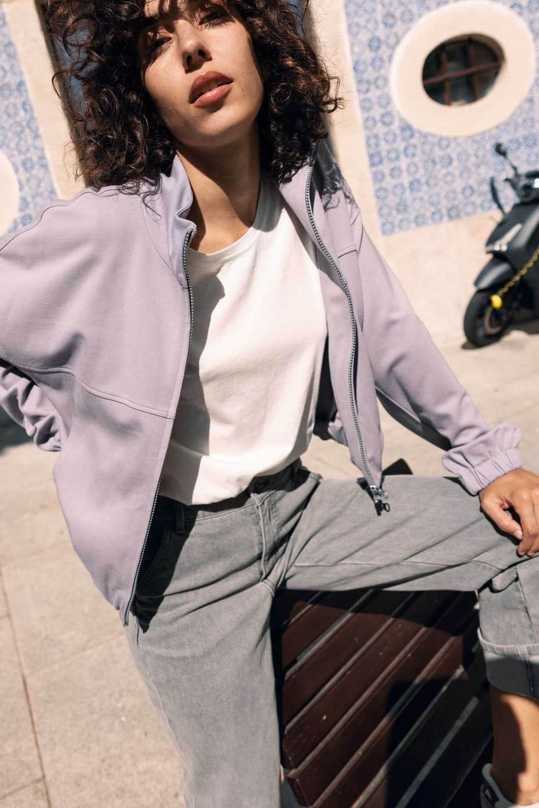 Femme portant une veste en sweat avec fermeture éclair de couleur lavande, assise en extérieur.