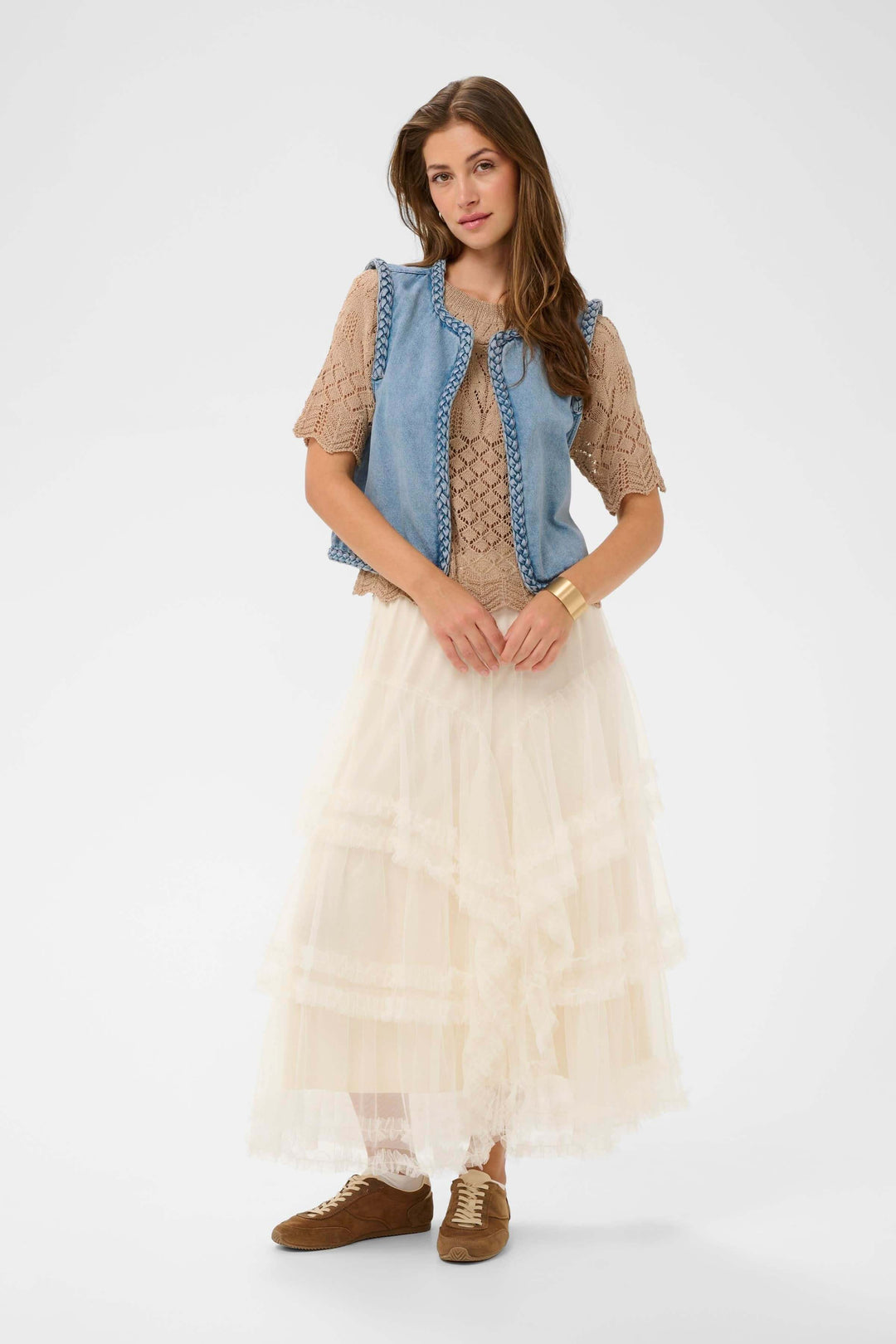 Femme portant la Veste sans manches Rina en denim avec bordure tressée, style boho chic avec jupe longue et blouse légère.