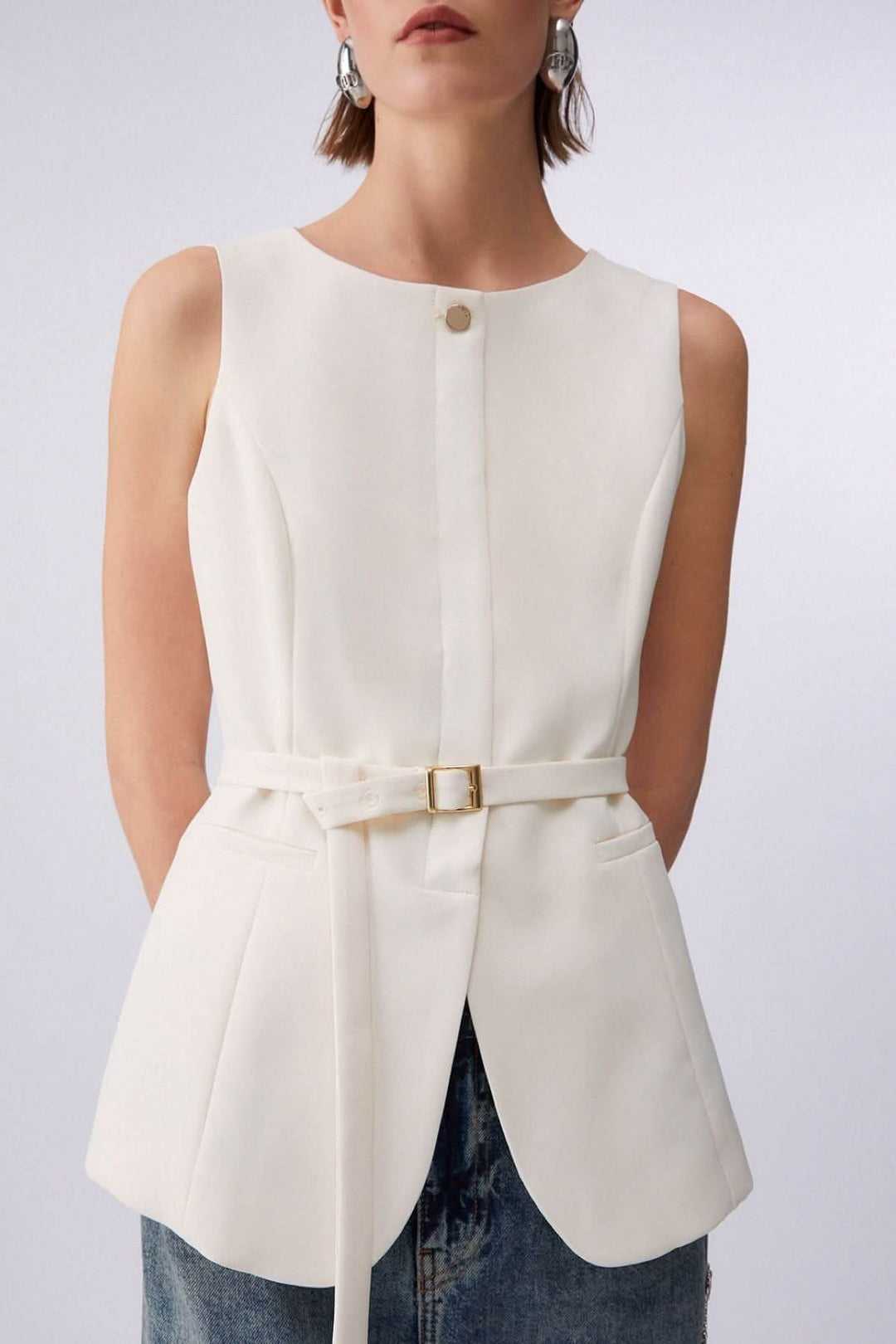 Veste sans manches avec ceinture blanche, bouton décoratif, encolure ronde, portée par une femme, design élégant et structuré.