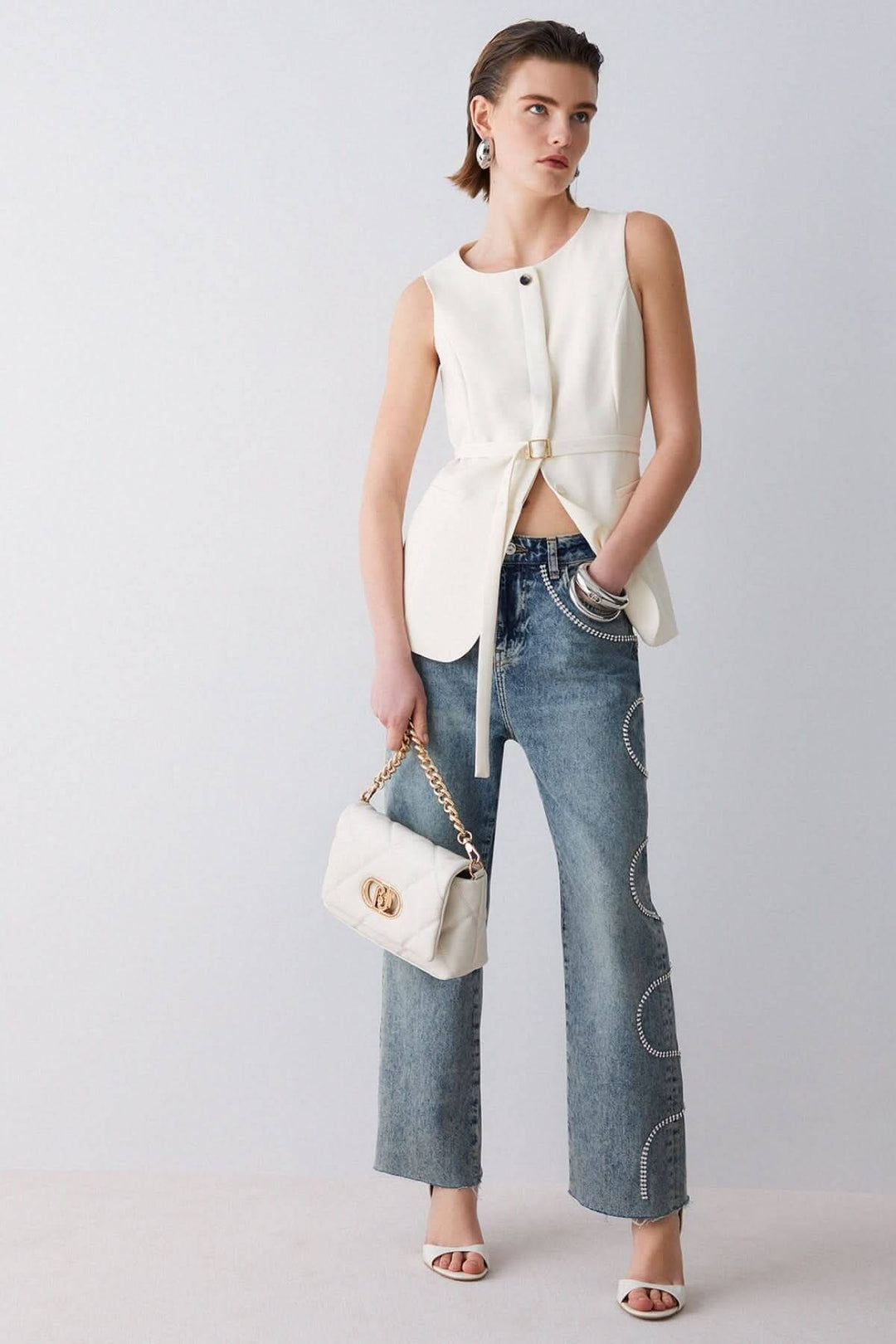 Veste sans manches avec ceinture, femme portant une tenue moderne et élégante avec jean et sac à main blanc, encolure ronde.