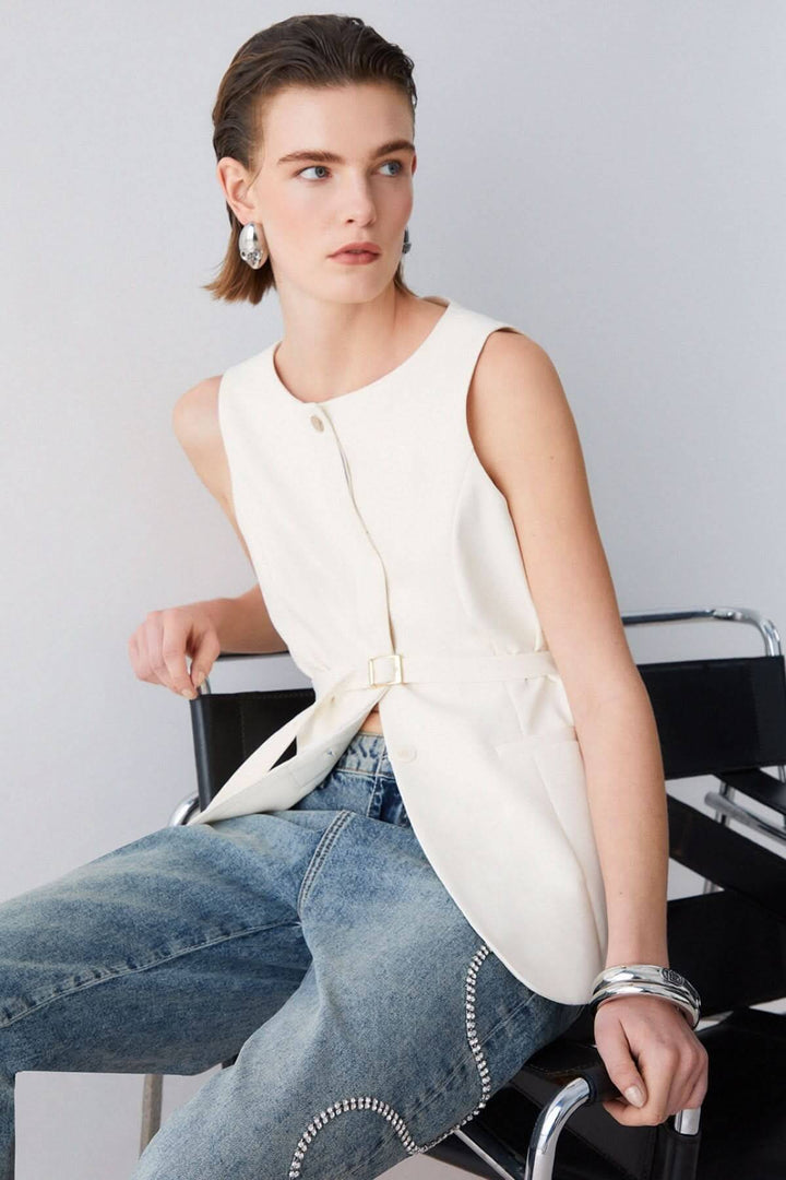 Femme portant une veste sans manches avec ceinture, assise sur un fauteuil, look moderne et sophistiqué.