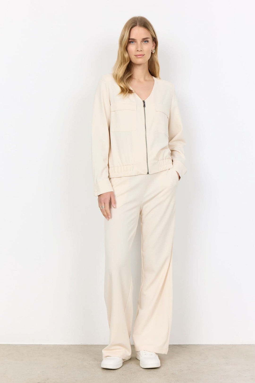 Veste zipée beige avec col en V, portée avec un pantalon assorti, idéale pour tenues décontractées ou professionnelles.