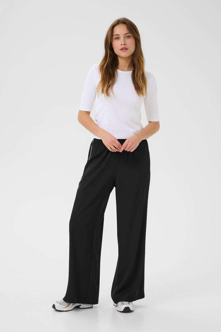 Femme portant le Pantalon Tessa fluide à jambe large, associé à un t-shirt blanc avec des baskets noires.