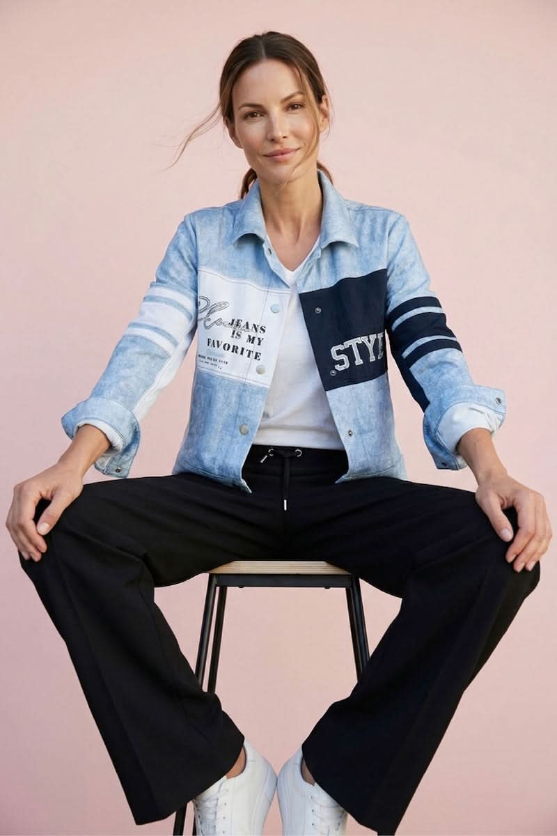 Femme assise sur un tabouret portant un pantalon jogging fluide noir et une veste en jean moderne, sur fond rose.