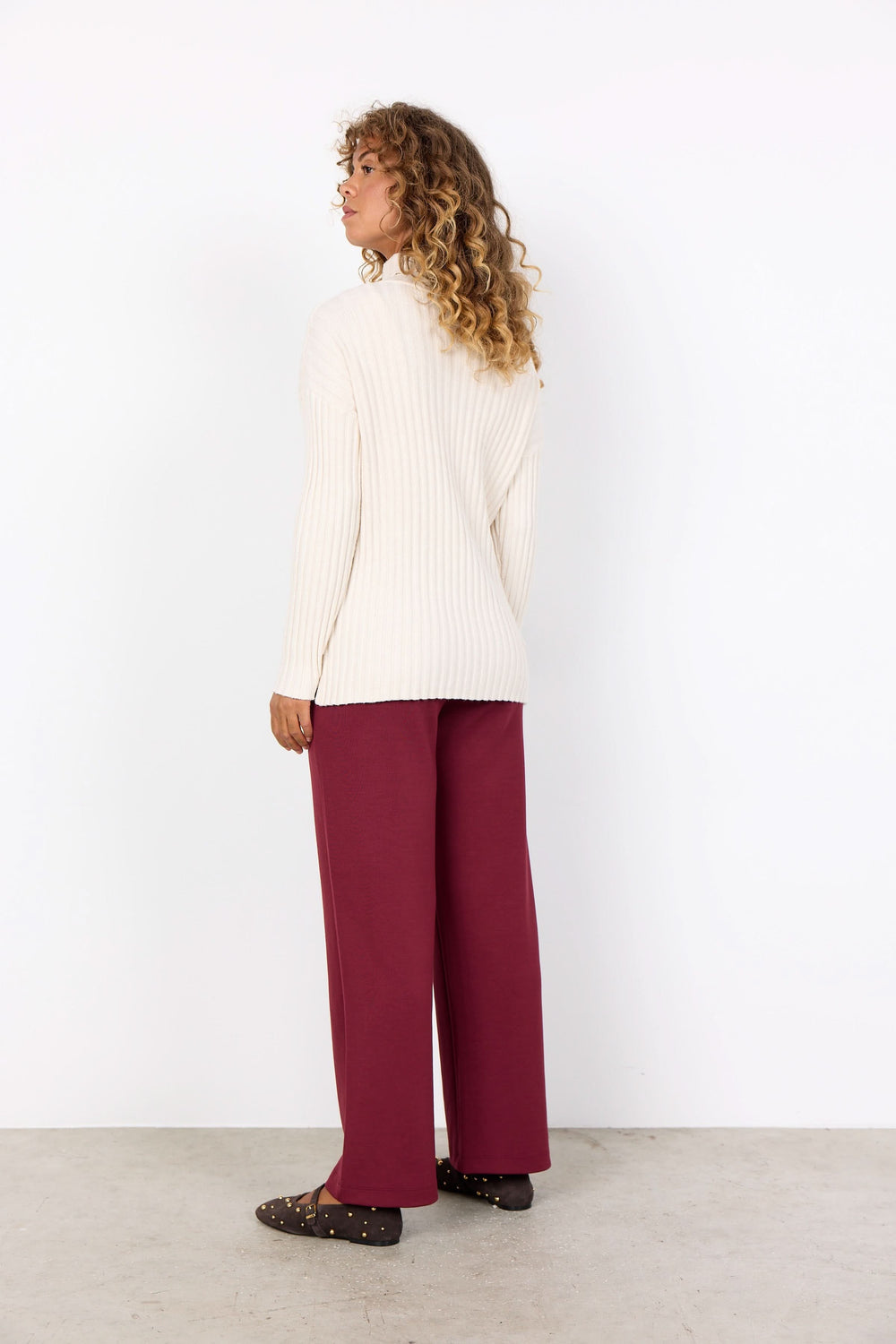 Femme vue de dos portant un tricot col montant crème et un pantalon ample bordeaux, ambiance décontractée chic.
