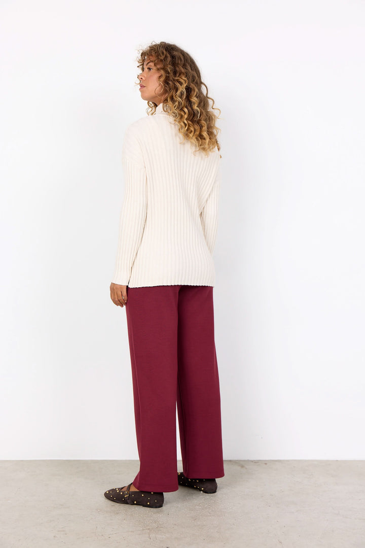 Femme vue de dos portant un tricot col montant crème et un pantalon ample bordeaux, ambiance décontractée chic.