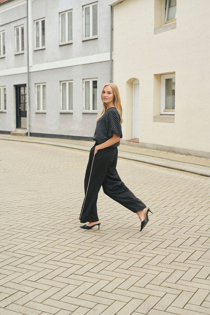 Femme marchant dans un pantalon fluide noir à jambe large et un haut à pois, dans une rue pavée.