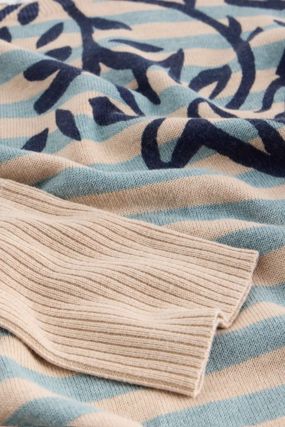 Chandail imprimé avec motif graphique, coupe droite, encolure ronde, et manches longues, sur fond rayé beige et bleu.