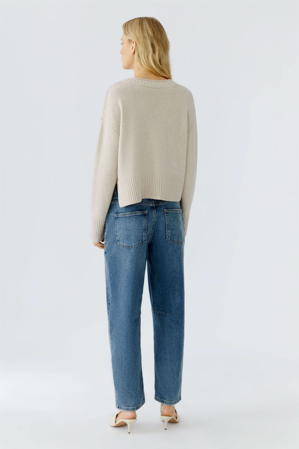 Femme de dos portant un chandail beige à poche, associé à un jean décontracté, style moderne et confortable.