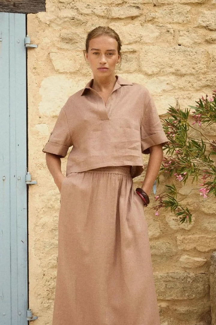 Femme portant une blouse courte en lin à col ouvert, élégante et décontractée, contre un mur en pierre, idéale pour l'été.