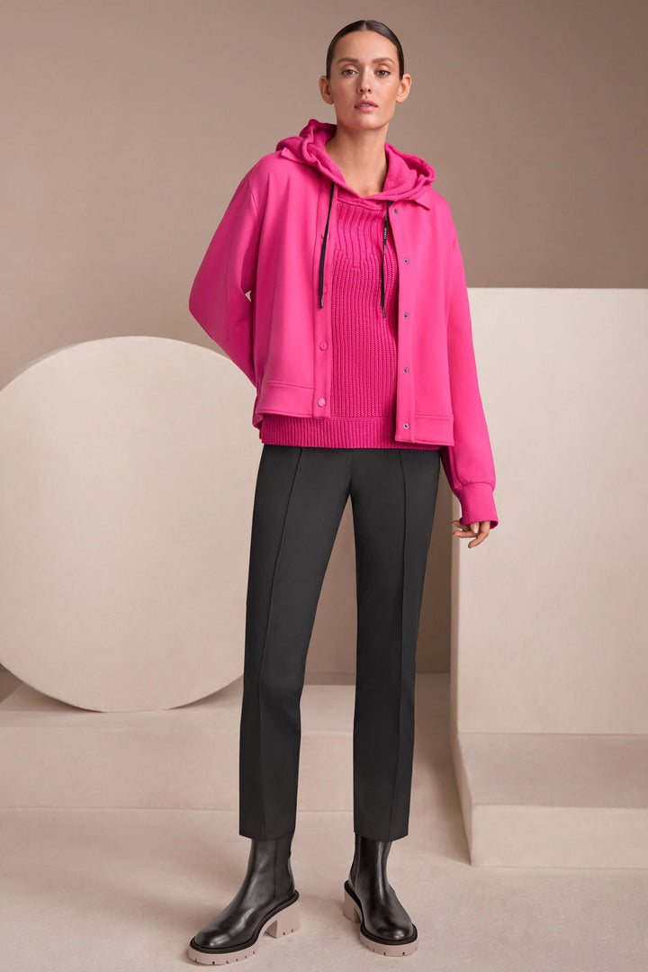 Femme en veste rose moderne avec pull à col montant et pantalon noir, pose décontractée sur fond neutre.