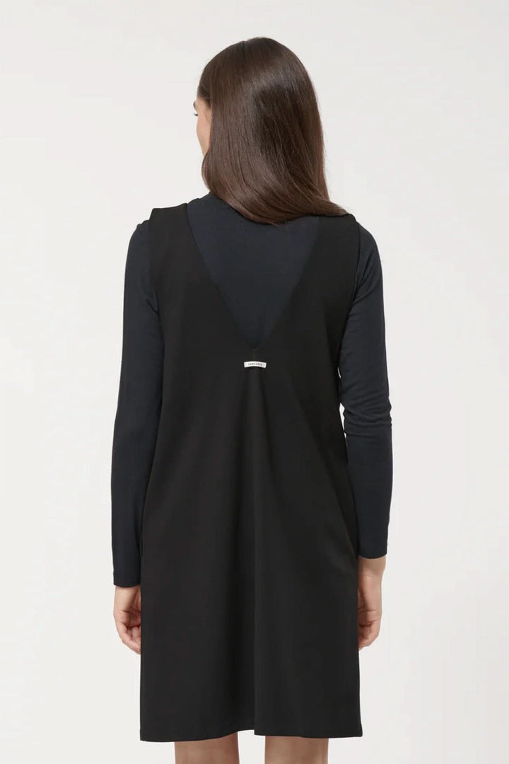 Femme portant une robe sans manches noire à encolure en V, coupe trapèze, sur un sous-pull noir.