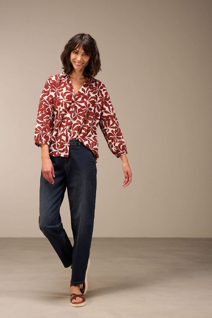 Femme souriante en chemisier floral rouge et jean décontracté, stylée et confortable pour un look moderne.