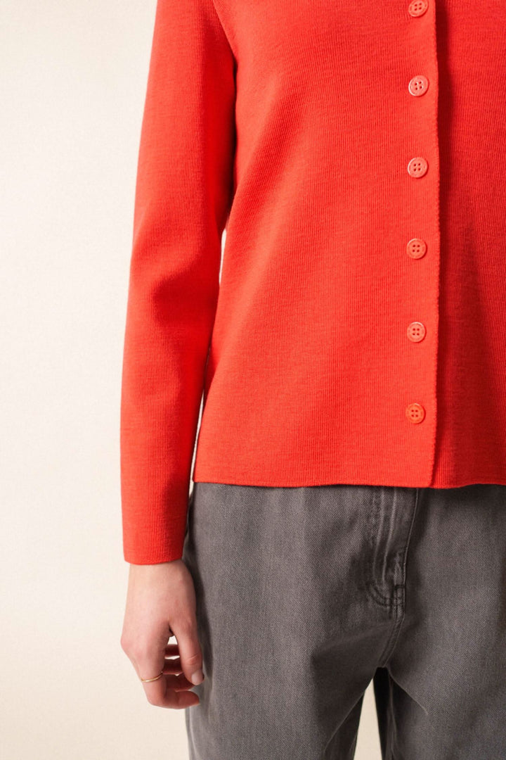 Gilet en laine douce à encolure en V, boutons discrets, couleur rouge, style minimaliste, porté avec un pantalon gris.