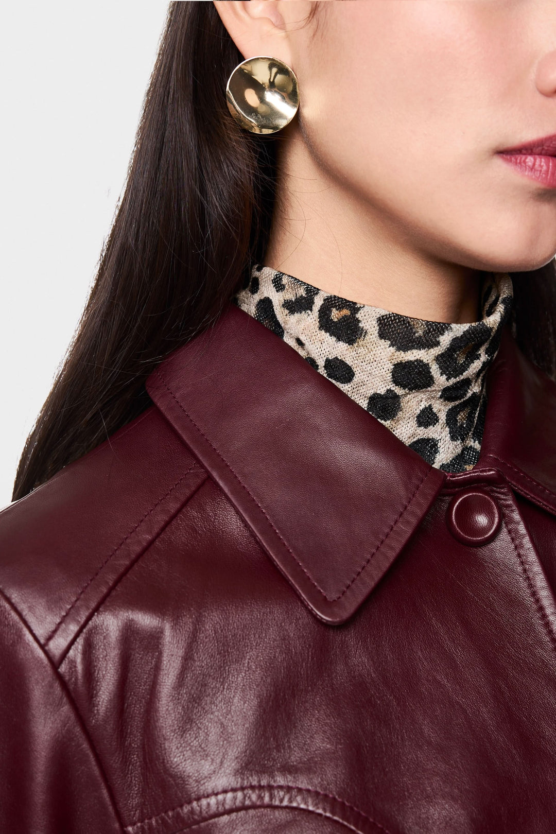 Détail d'une veste en cuir bordeaux portée avec un col roulé à motif léopard et des boucles d'oreilles dorées.