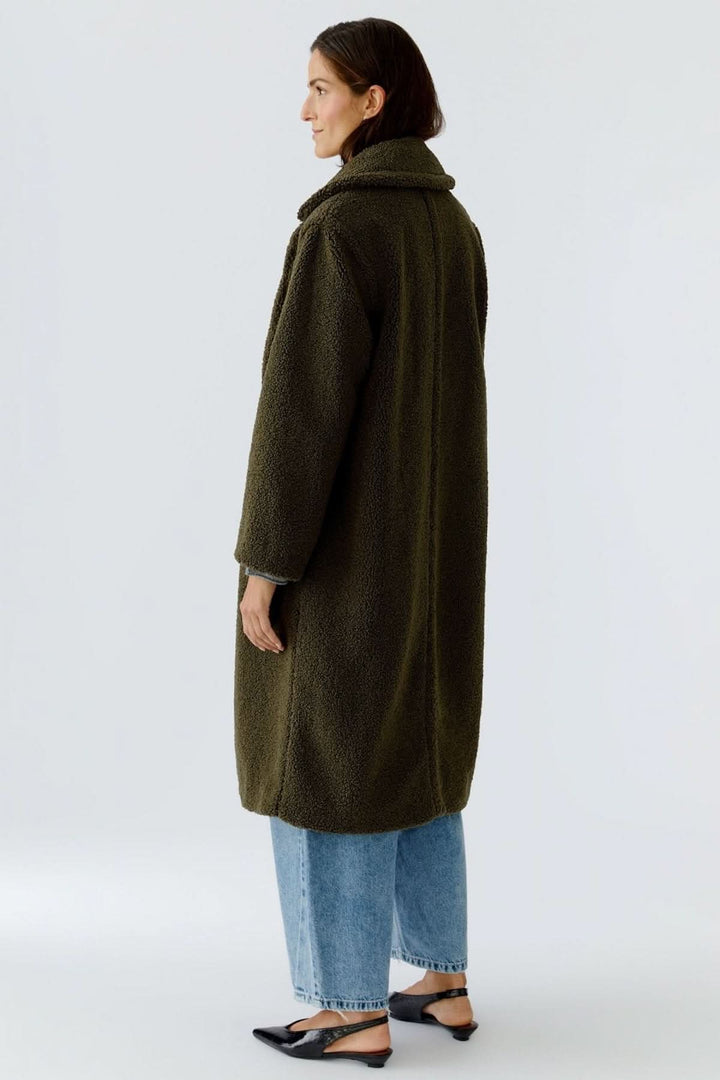 Manteau long en tissu bouclette vert foncé, porté par une femme, de dos, avec un jean et des chaussures noires.