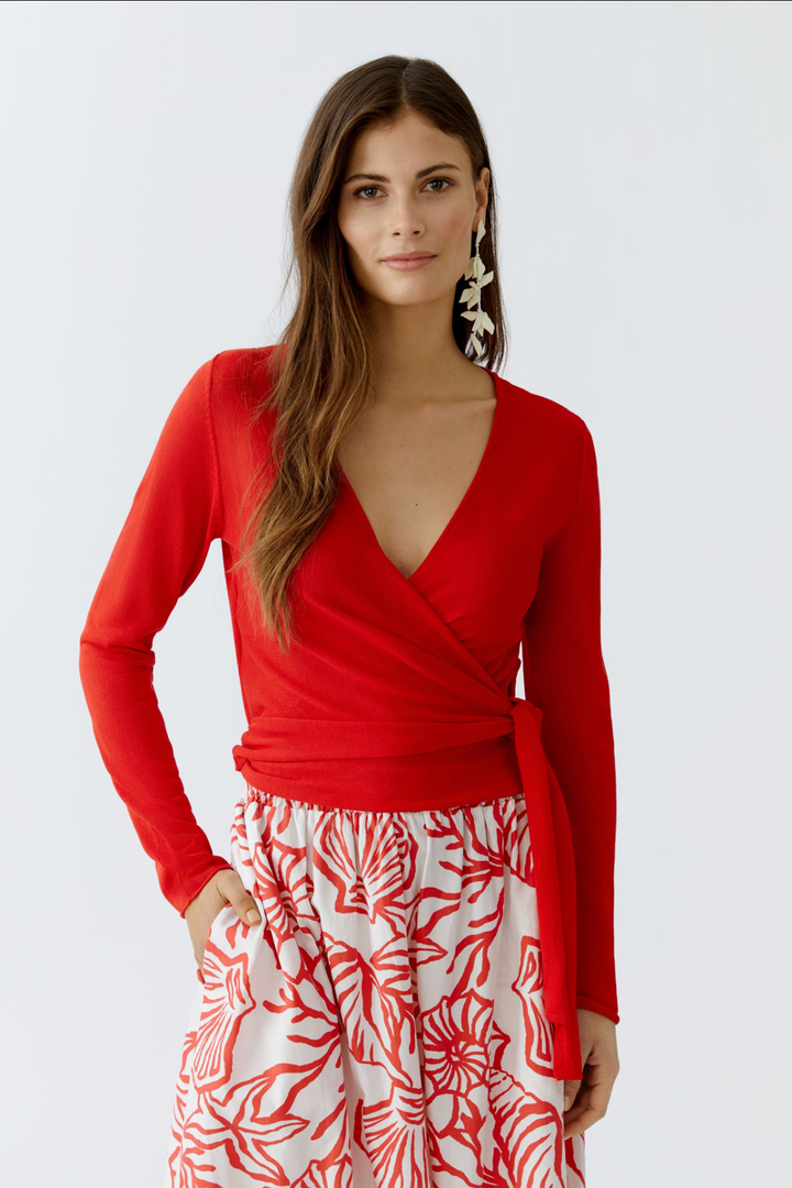 Chandail portefeuille rouge à décolleté en V, manches longues et lien à nouer, porté avec une jupe imprimée.