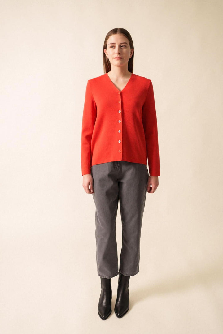 Gilet en laine rouge à encolure en V, boutons discrets, porté avec un pantalon gris et des bottines noires.