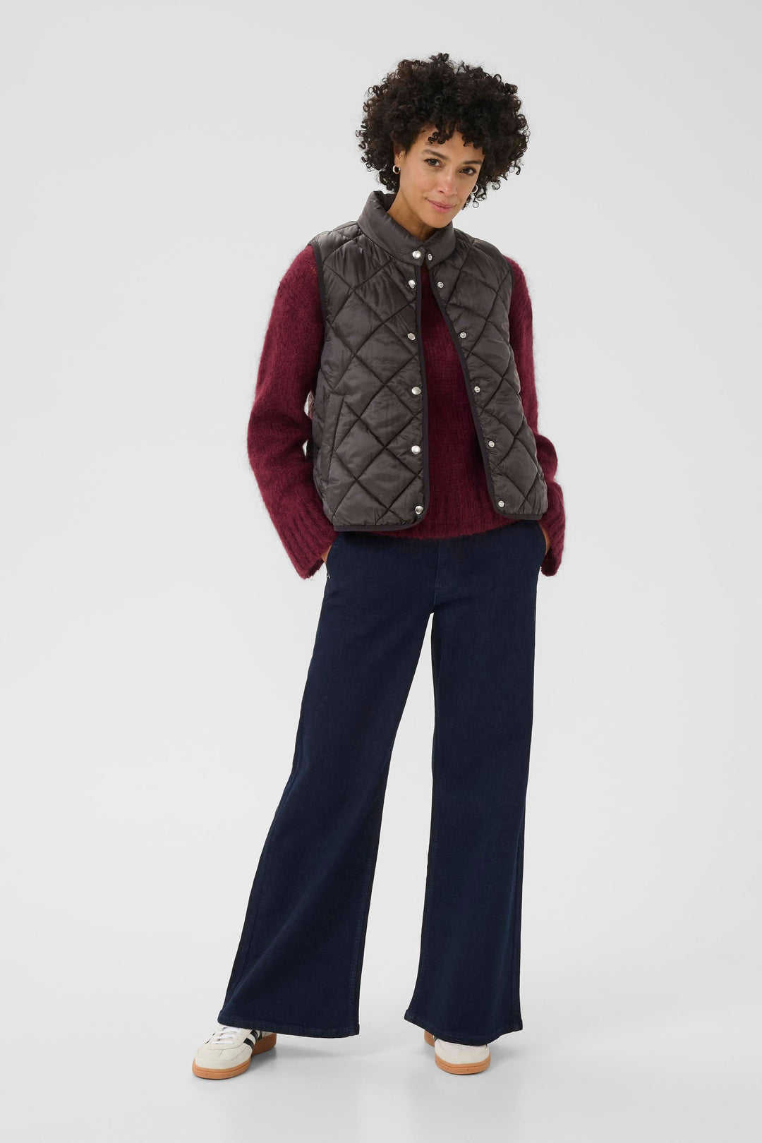 Manteau sans manches matelassé gris sur un chandail bordeaux, pantalon large en denim, look décontracté moderne.
